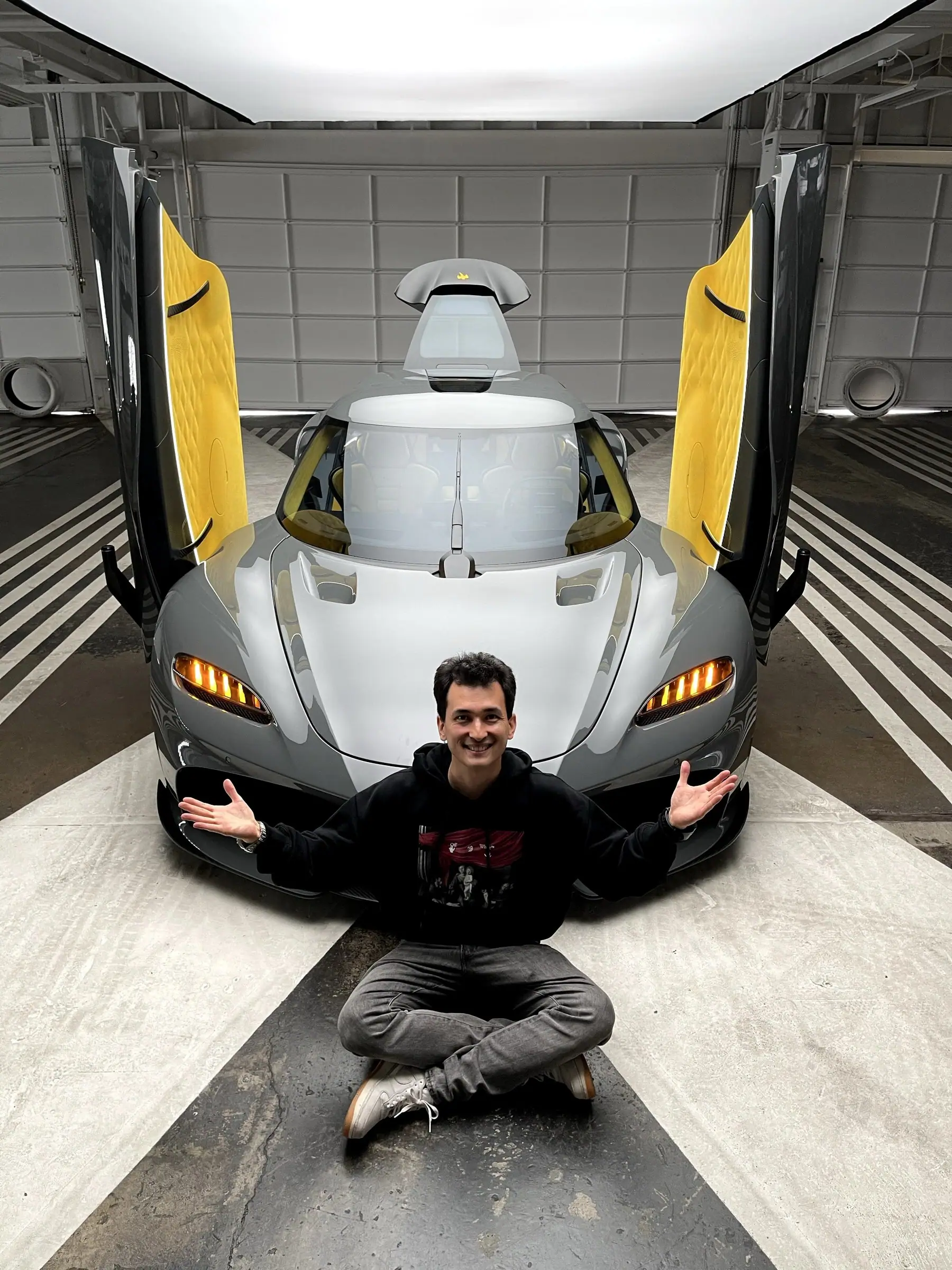 Alan sitting in front of a silver hypercar with doors raised in a studio
