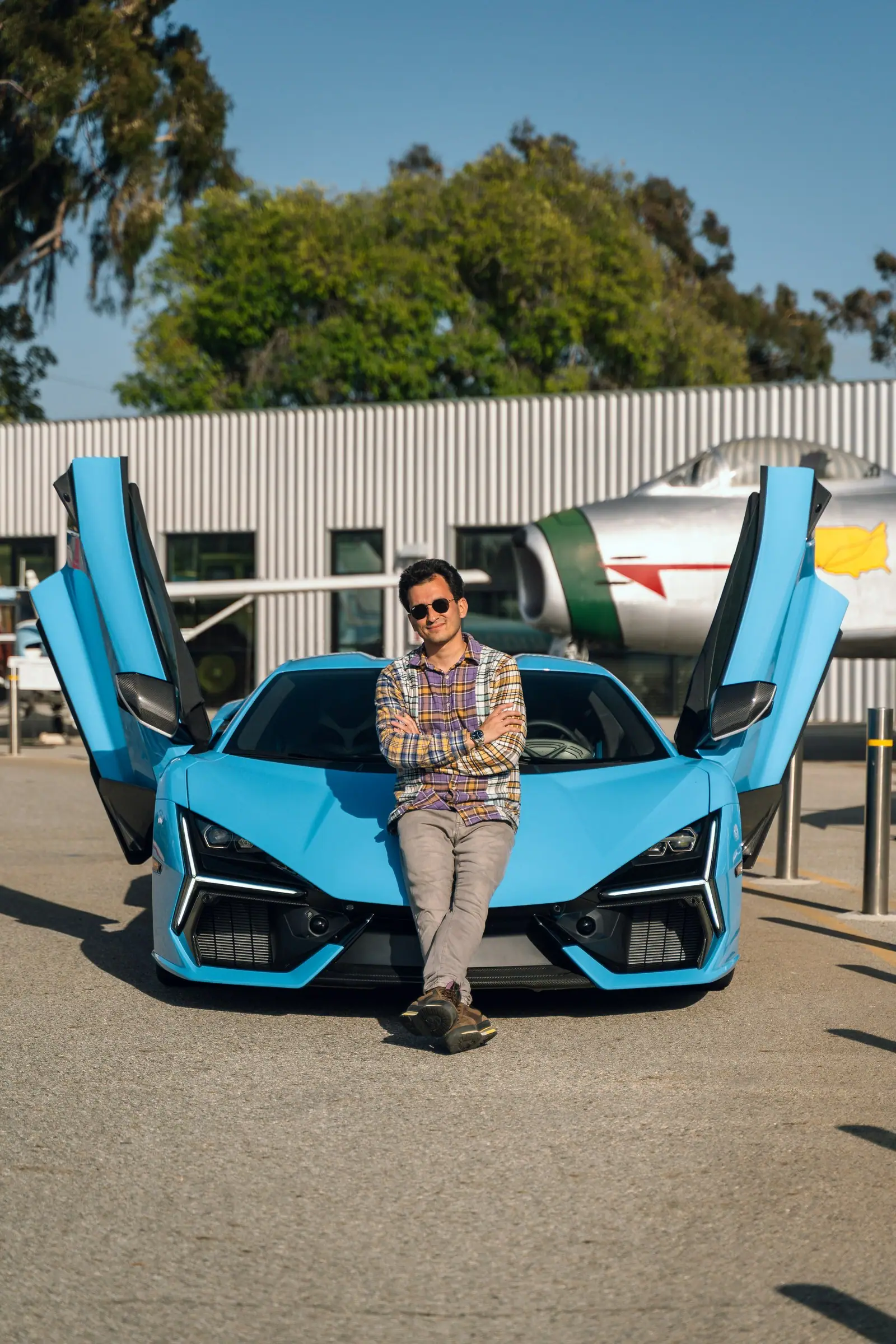 Alan seated on the nose of a blue Lamborghini Revuelto with both doors raised