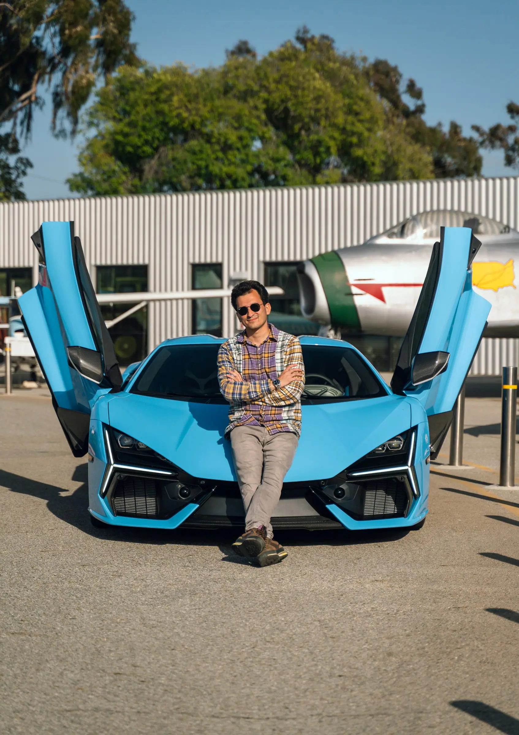 Alan Enileev seated on the nose of a blue Lamborghini Revuelto with both doors raised